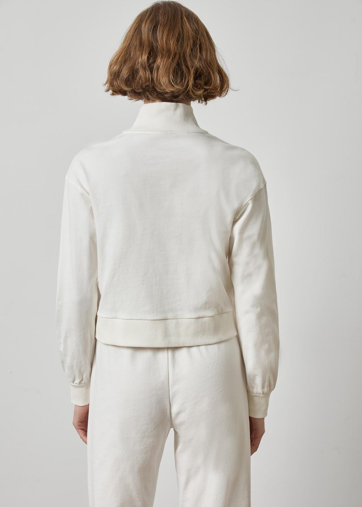 A model viewed from the back wearing a cream-colored long-sleeve cropped knit top and matching cream pants against a light gray studio background.