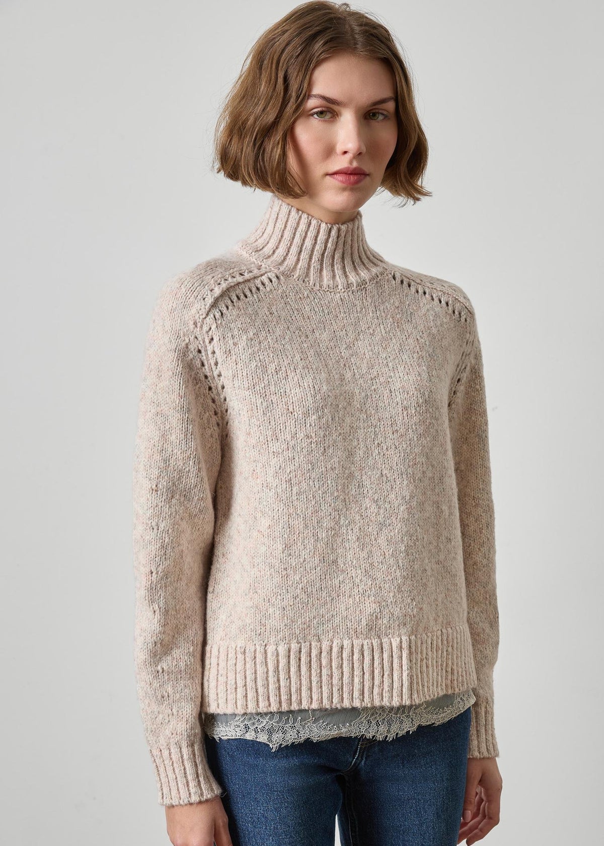 A woman wearing a beige knit mock neck sweater with textured ribbing and openwork details on the shoulders, paired with blue jeans, against a light gray studio background.