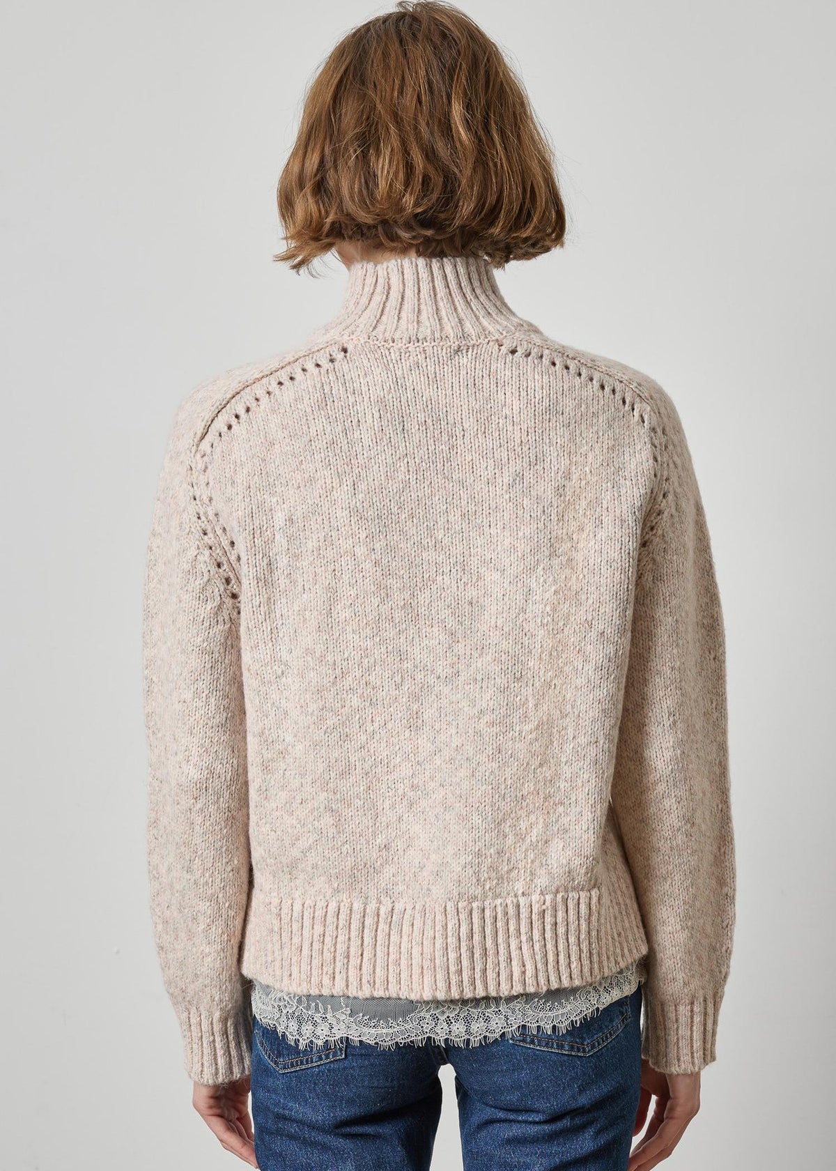 A person shown from the back wearing a light beige knit turtleneck sweater with textured knit details and ribbed hem and cuffs, paired with blue jeans, against a plain light gray studio background.