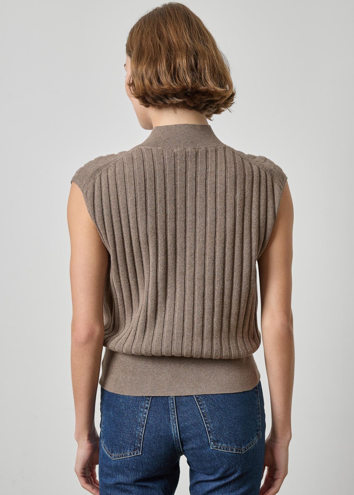 A woman viewed from the back wearing a sleeveless ribbed knit mock-neck sweater vest in taupe brown, paired with blue jeans, against a light gray studio background.