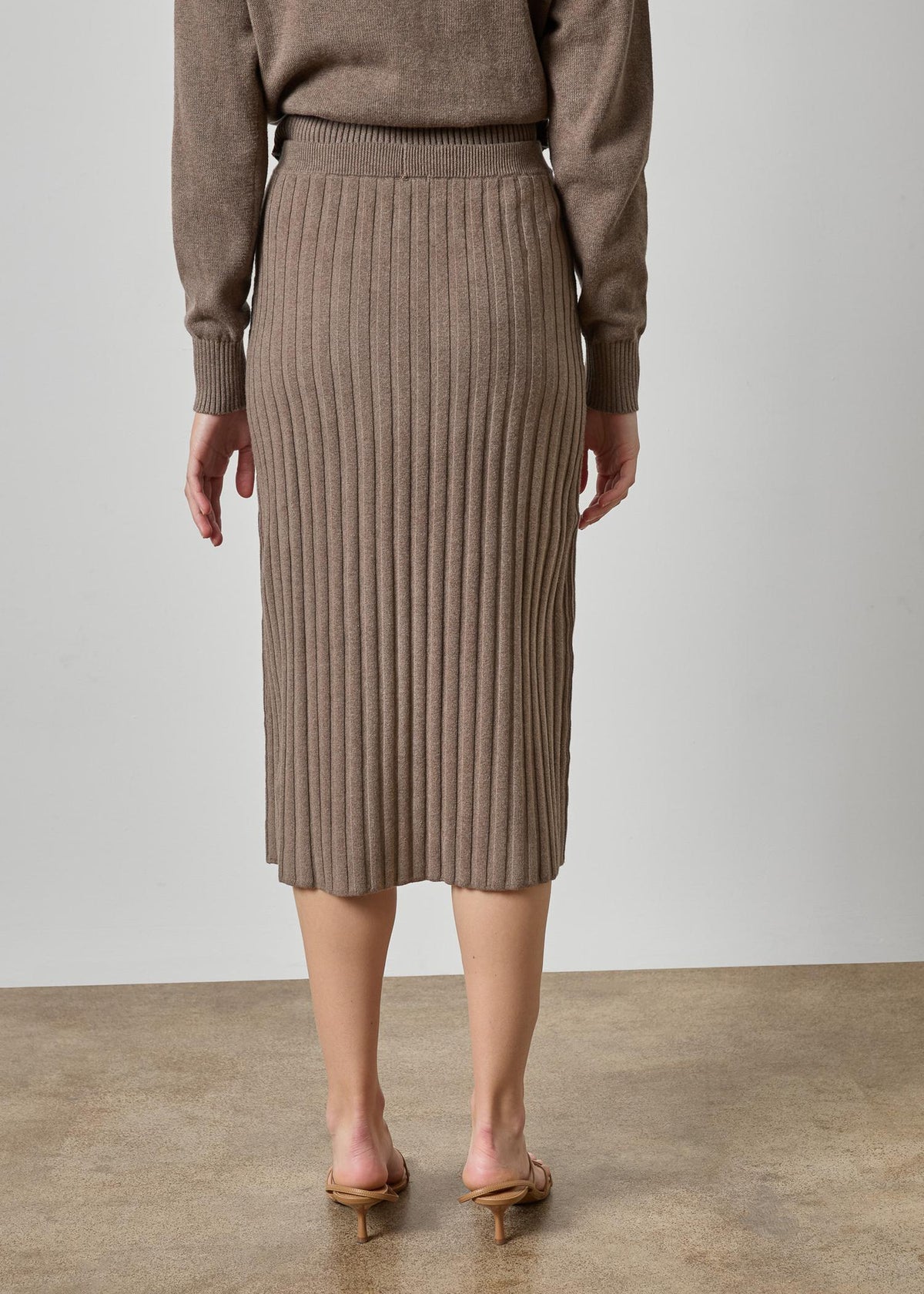 A model from the back wearing a taupe brown ribbed knit midi skirt and matching sweater, paired with tan heeled sandals, set against a neutral studio background.