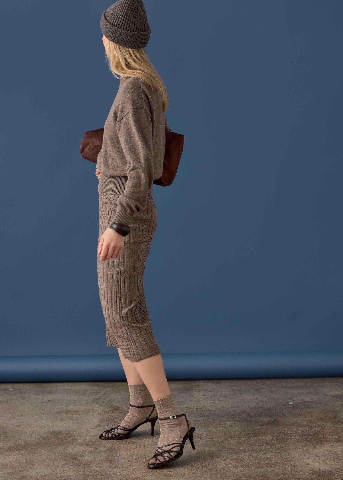 A woman stands in profile wearing a ribbed taupe knit sweater and matching ribbed midi skirt, a dark gray beanie, beige socks, black strappy heels, and carrying a brown shoulder bag against a blue studio backdrop.