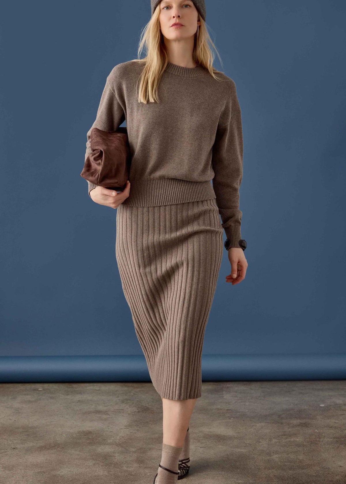 A woman models a taupe brown ribbed knit sweater and matching midi skirt set, paired with a matching beanie, a brown leather clutch, and ankle boots, set against a solid blue studio backdrop.