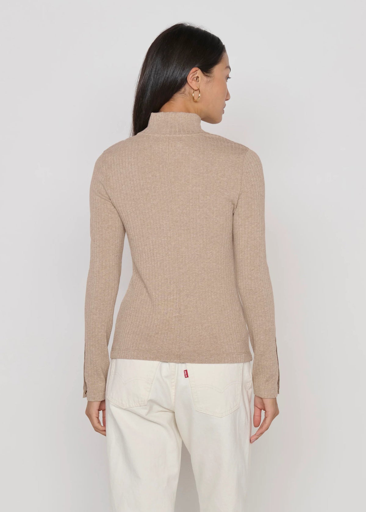 A woman shown from the back wearing a beige ribbed mock neck sweater and white trousers, photographed against a light gray studio background.