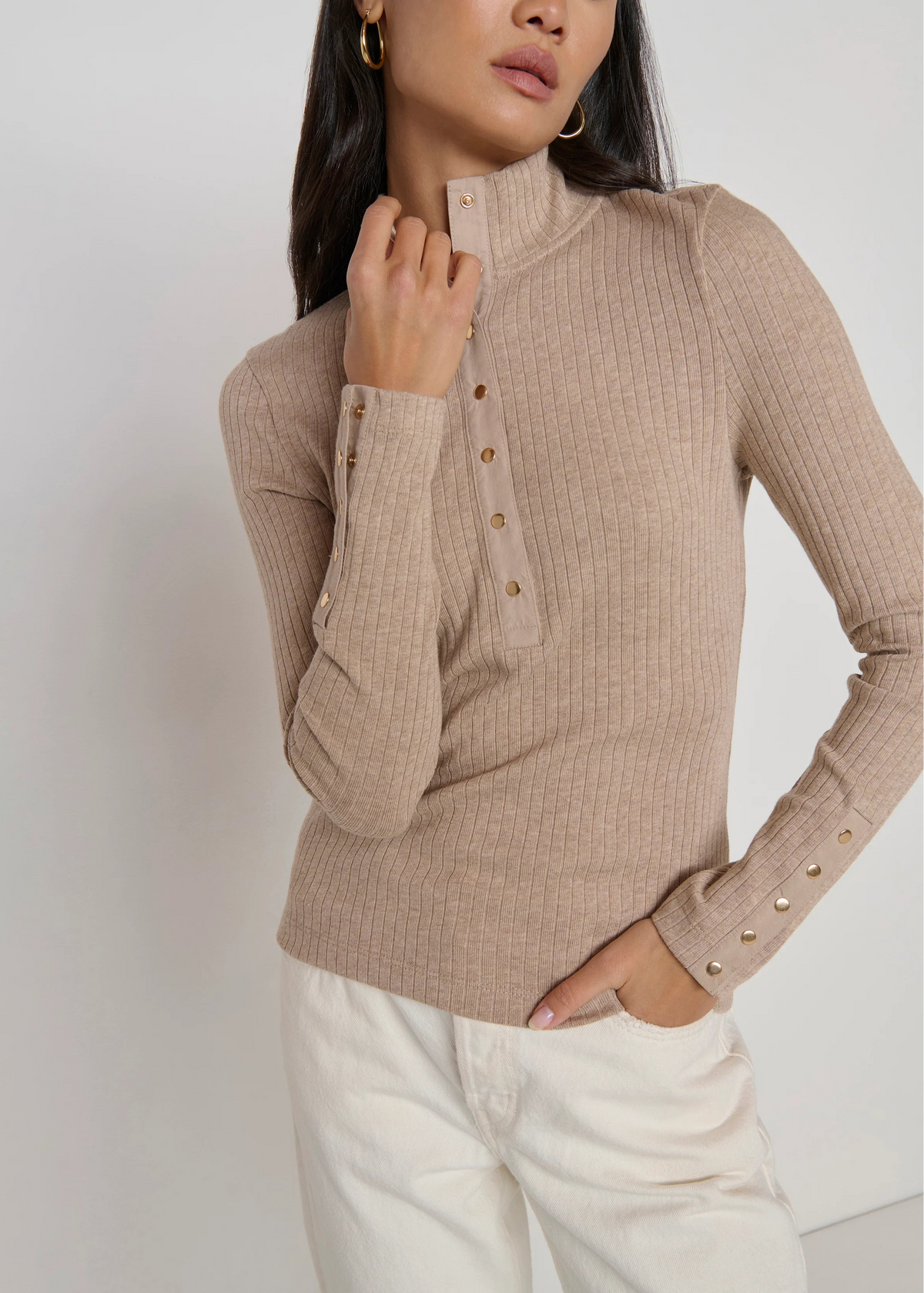 A woman wearing a beige ribbed mock-neck long-sleeve top with gold-tone decorative buttons down the front and cuffs, paired with white pants.