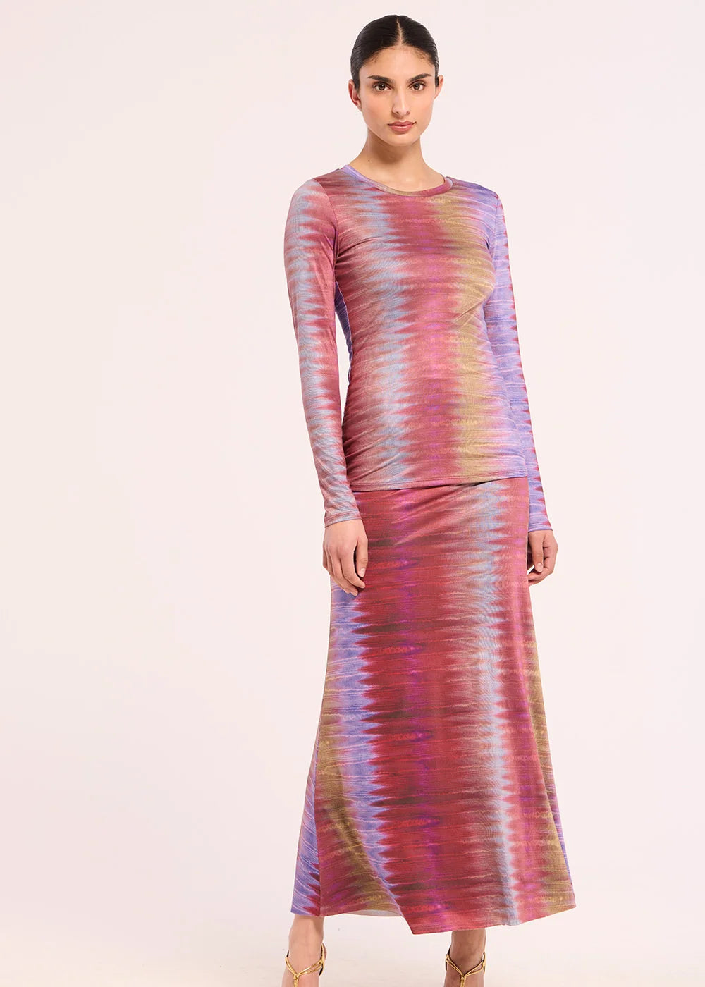 A woman modeling a long-sleeve maxi dress with a vibrant, wavy multicolor tie-dye pattern in pink, purple, blue, and yellow, paired with gold strappy heels against a plain light background.