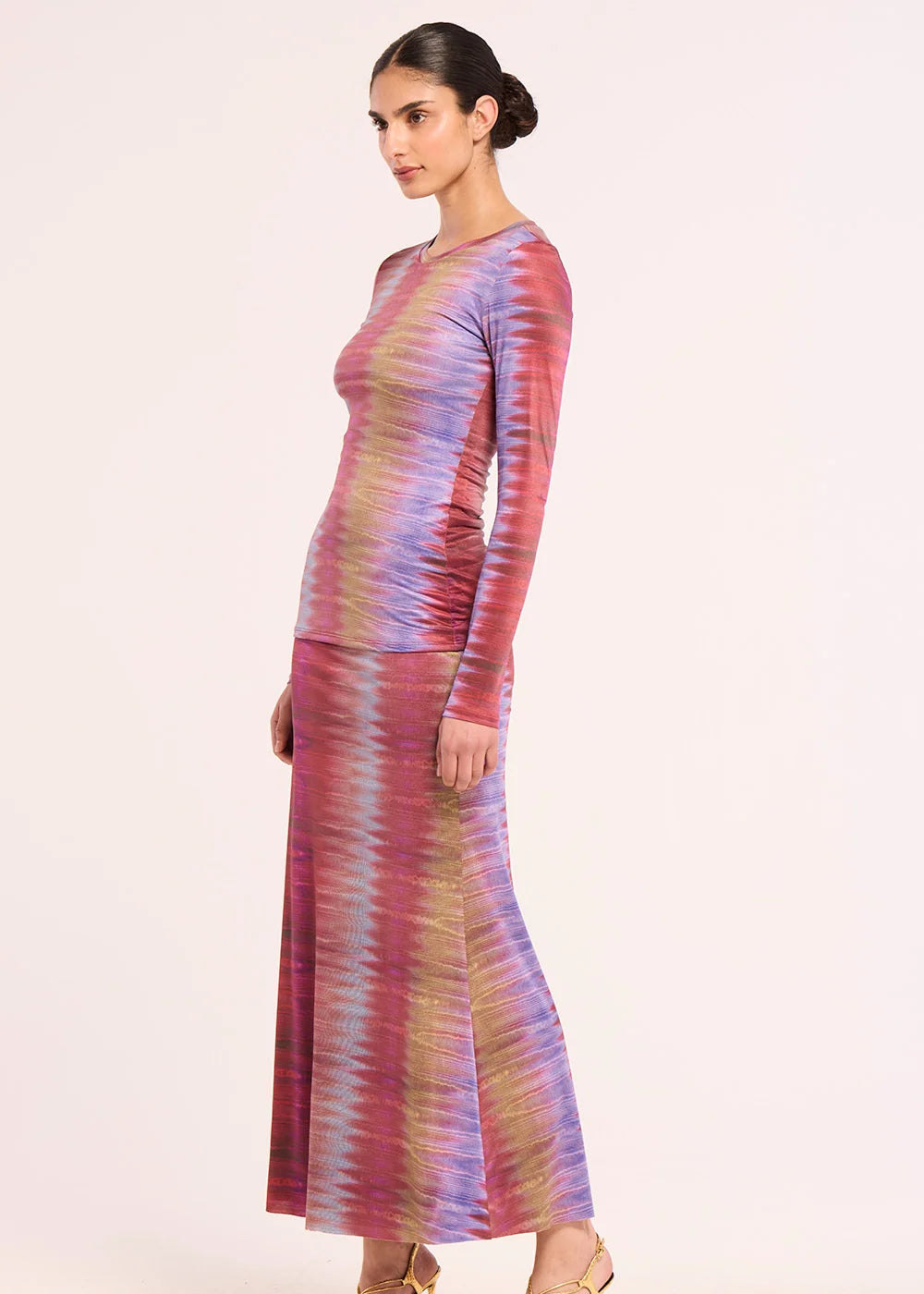 A woman modeling a long-sleeve maxi dress with a vertical wavy stripe pattern in purple, pink, and gold, shown from a side profile against a plain light background.