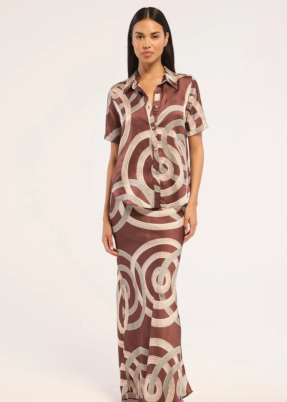 A woman modeling a matching two-piece lounge set consisting of a short-sleeve button-up shirt and a long maxi skirt, both featuring a brown and beige abstract circular pattern, set against a plain white studio background.