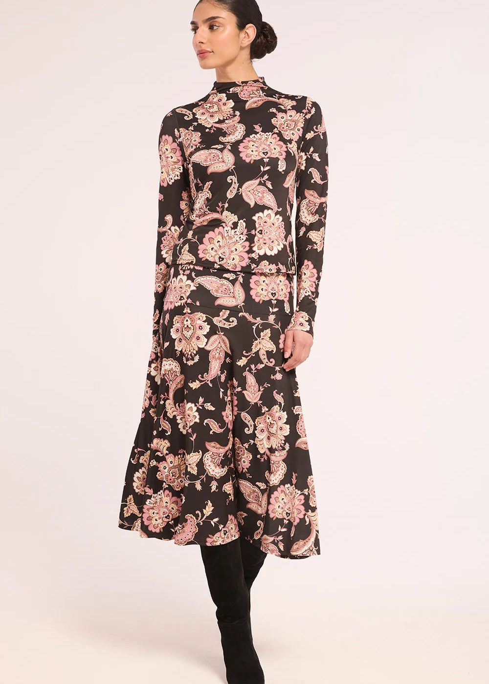 A woman modeling a long-sleeved midi dress with a black base and a pink floral paisley pattern, paired with black boots, against a plain light background.
