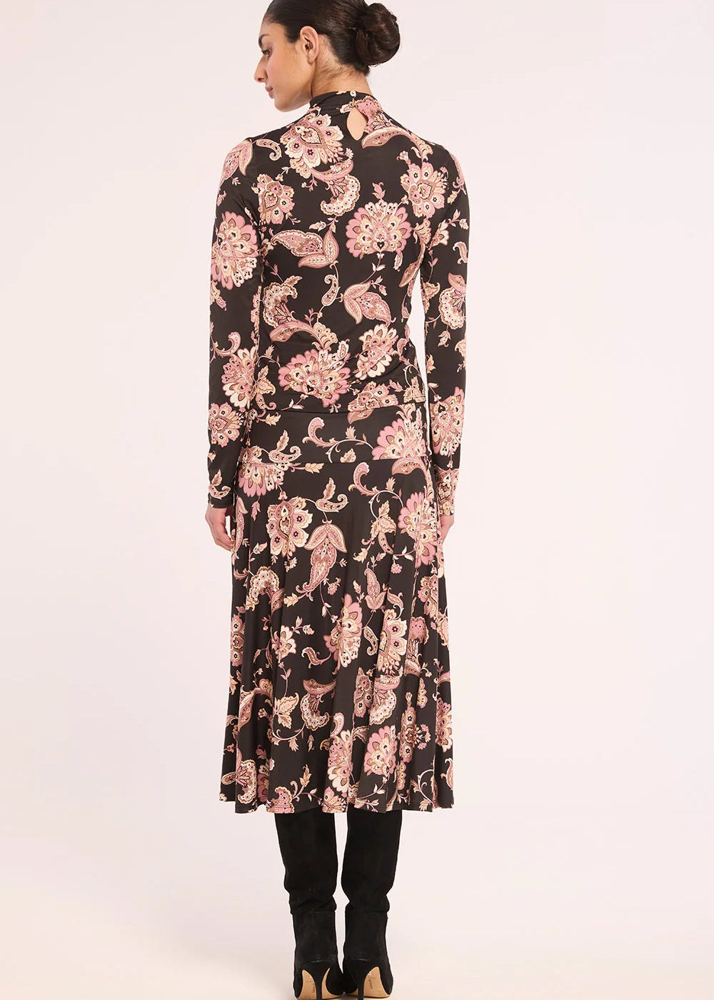 A woman viewed from the back wearing a long-sleeve midi dress with a black base and a pink floral pattern, paired with black boots, against a plain light background.