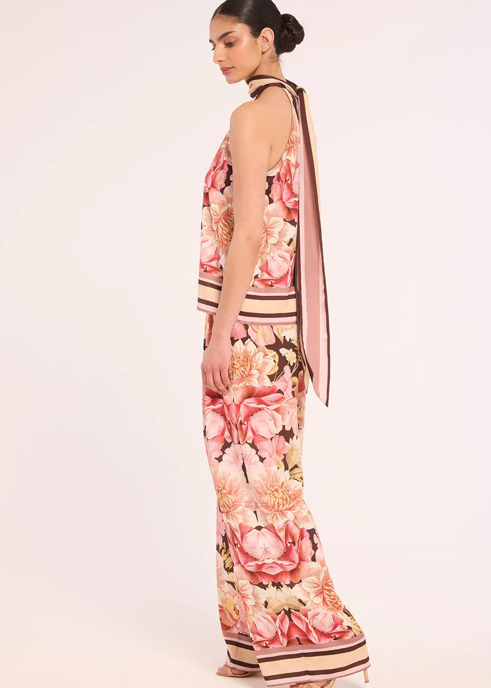 A woman modeling a sleeveless halter-neck maxi dress with a pink, peach, and beige floral print and striped border details, shown from a side-back angle against a plain light background.
