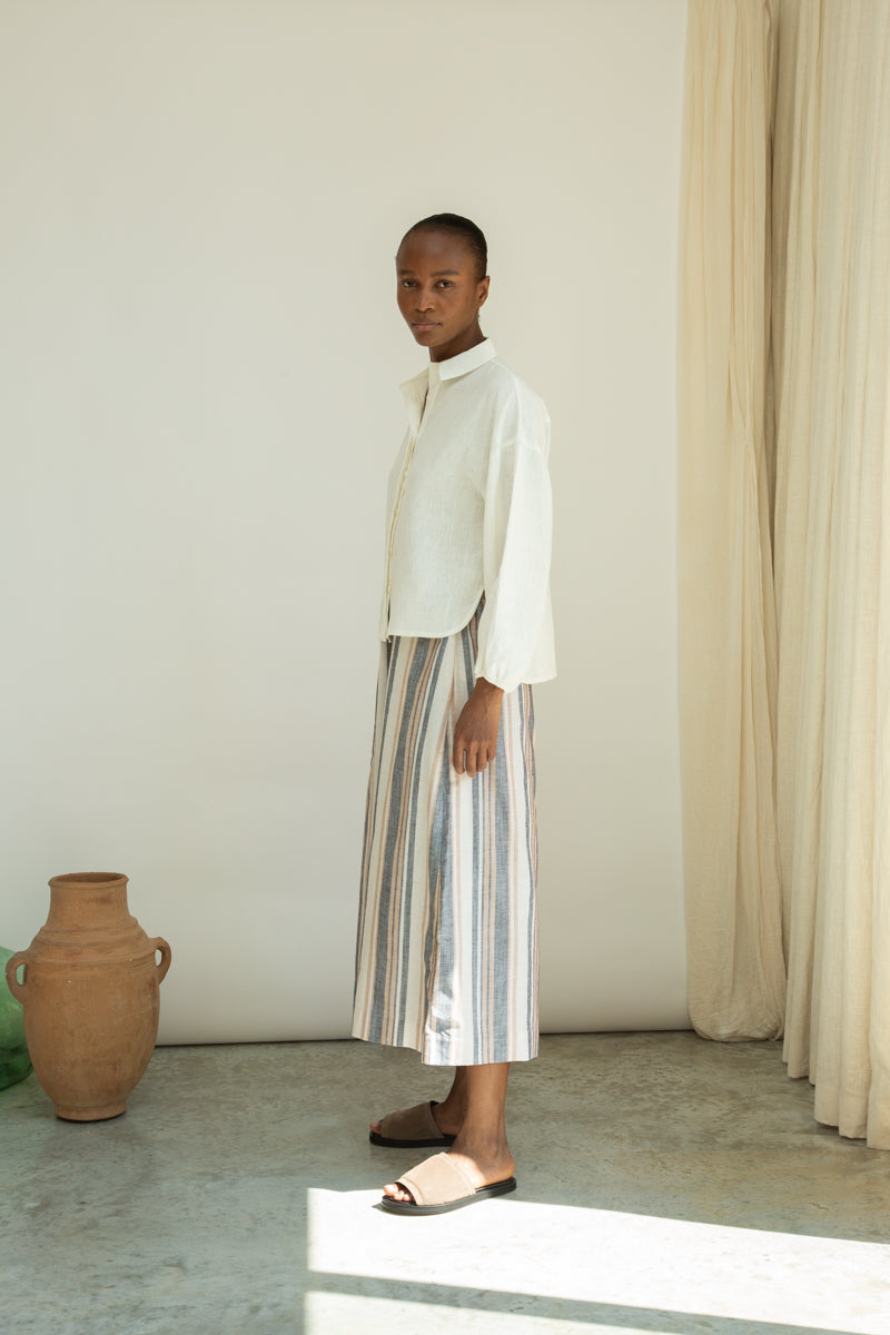 Woman in a light outfit standing in a minimalistic room with a vase and curtain.
