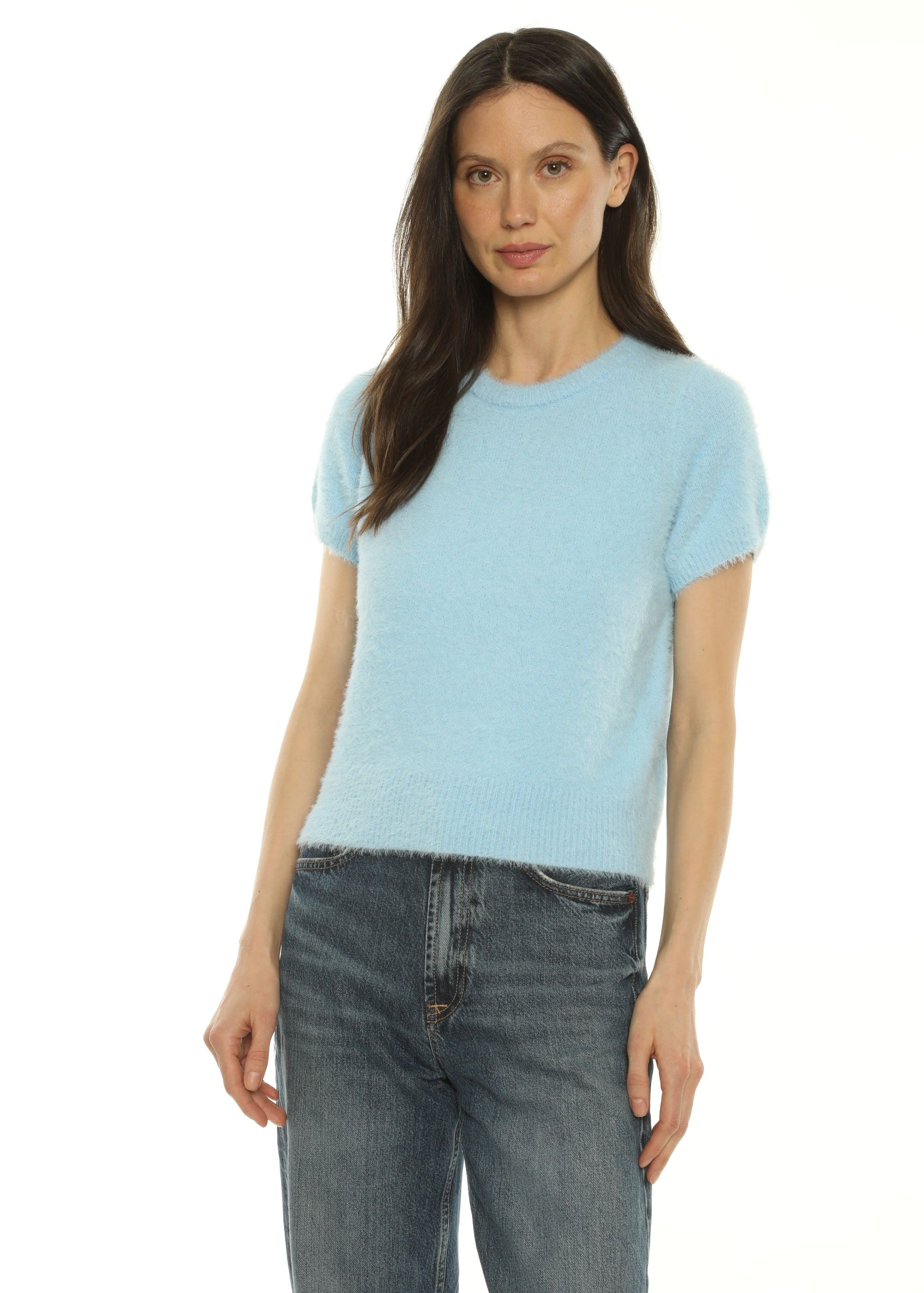 A woman wearing a light blue short-sleeve knit top and dark blue jeans, posed diagonally against a plain white background.