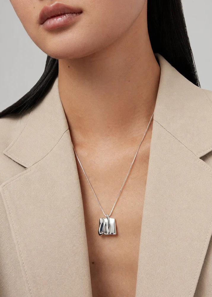 A close-up of a model wearing a silver pendant necklace with a textured, abstract block design, styled with a beige blazer.