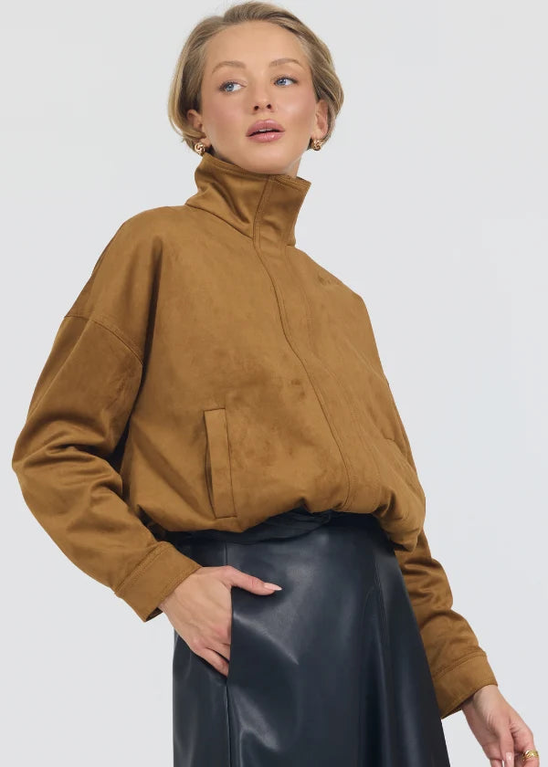 A woman wearing a camel brown suede-like high-neck jacket with a front zipper and side pockets, paired with a black faux-leather skirt, against a light gray studio background.