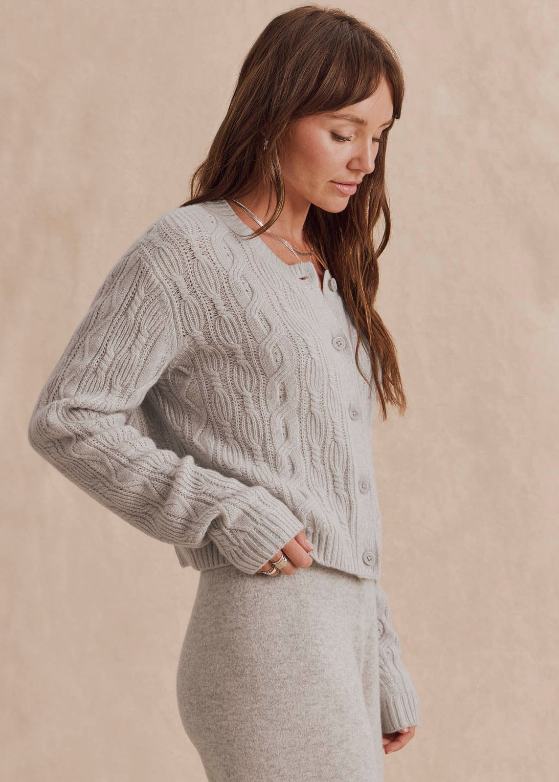 A woman wearing a light gray cable-knit cardigan sweater and matching knit skirt, posed against a warm beige textured studio backdrop.