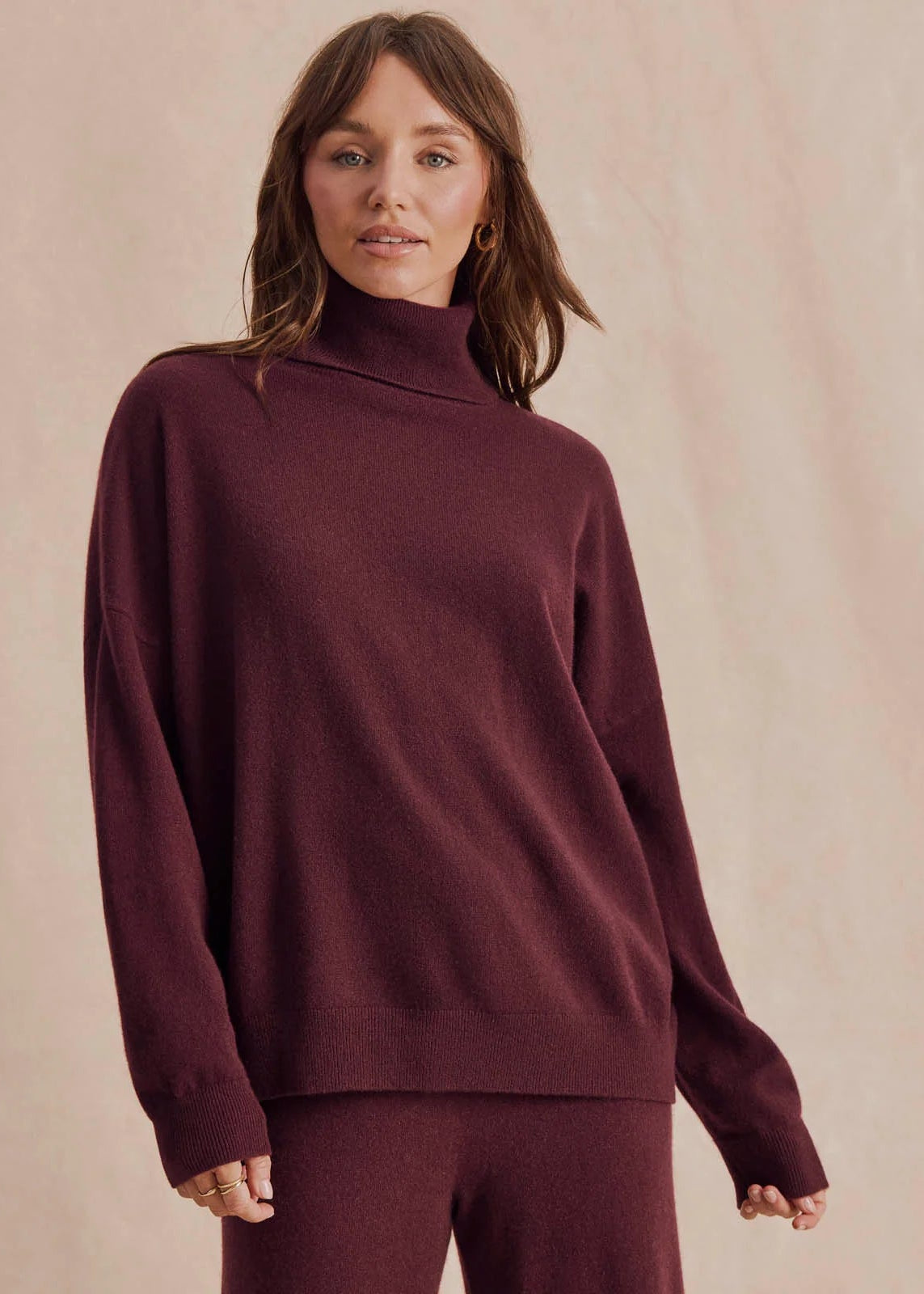 A woman wearing a matching burgundy knit turtleneck sweater and trousers, standing against a soft beige textured background.