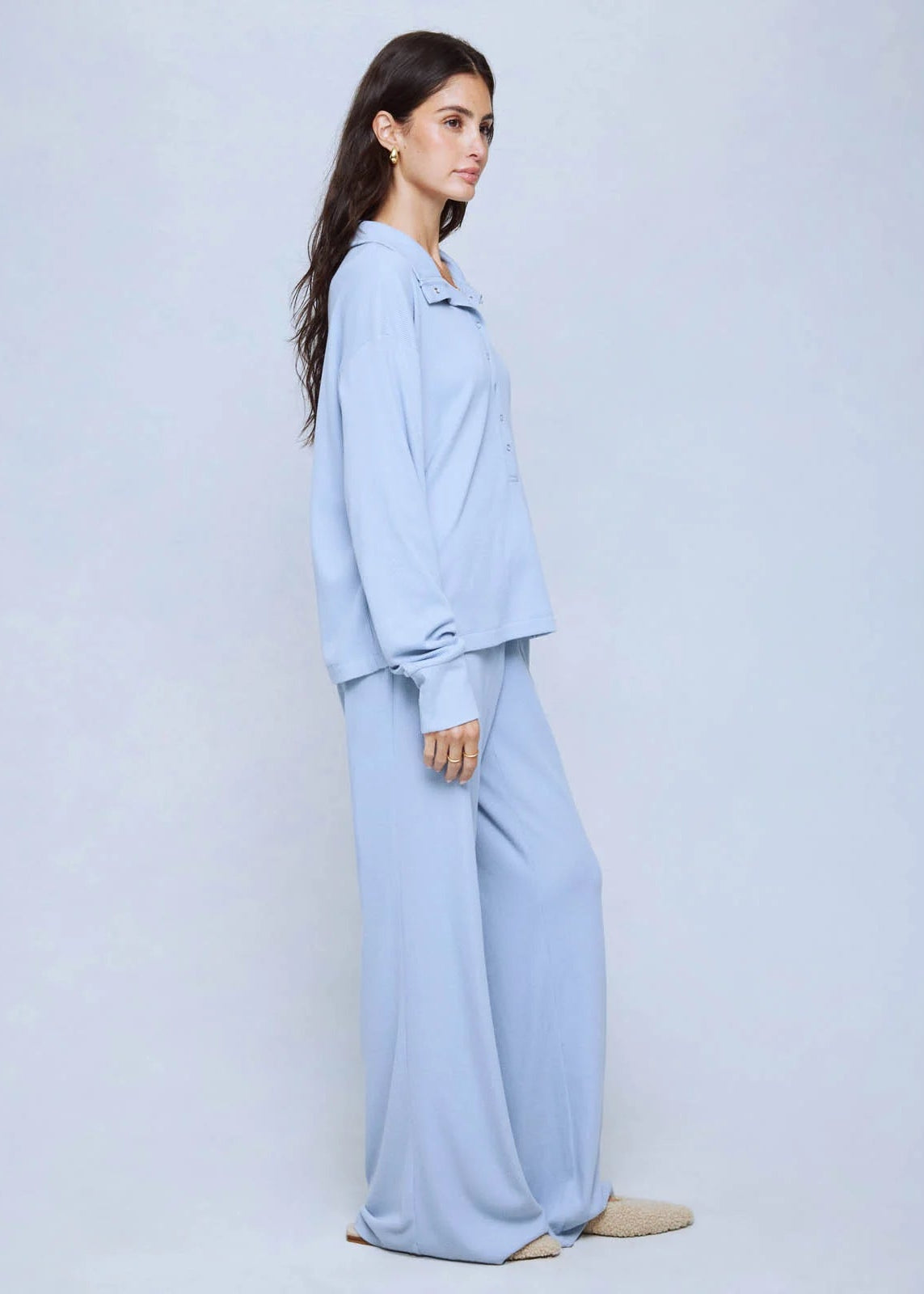 A woman modeling a light blue two-piece lounge set consisting of a long-sleeve button-up shirt and matching wide-leg pants, photographed against a solid light gray studio background.