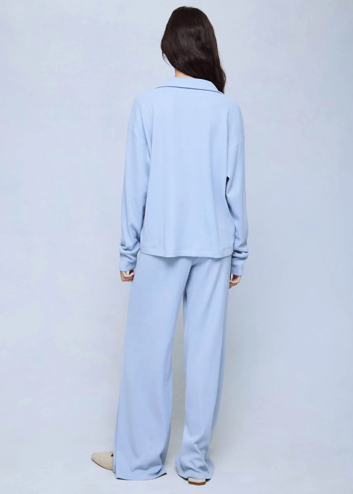 A woman viewed from behind wearing a light blue matching lounge set consisting of a long-sleeve collared top and wide-leg pants, standing against a plain light gray studio background.