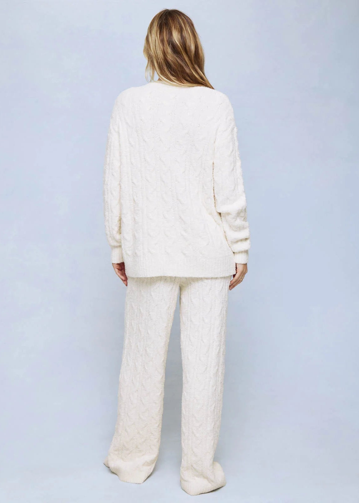 A woman viewed from behind wearing a matching cream cable-knit lounge set, consisting of a long-sleeve top and wide-leg pants, against a light blue studio background.