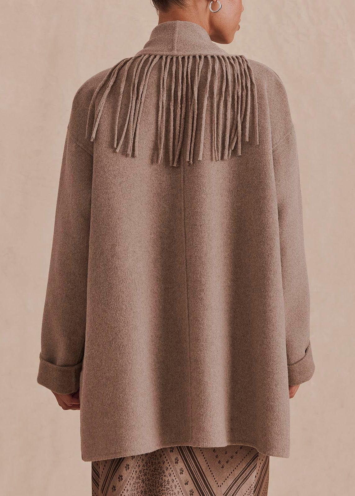 A person shown from the back wearing a taupe-brown long-sleeve coat with a high mock neck and a prominent fringe detail across the shoulders, paired with patterned pants against a textured beige backdrop.