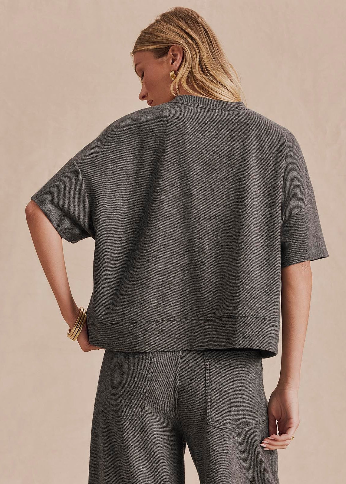 A woman shown from the back wearing a dark gray short-sleeve cropped sweatshirt and matching gray pants against a warm beige studio background.