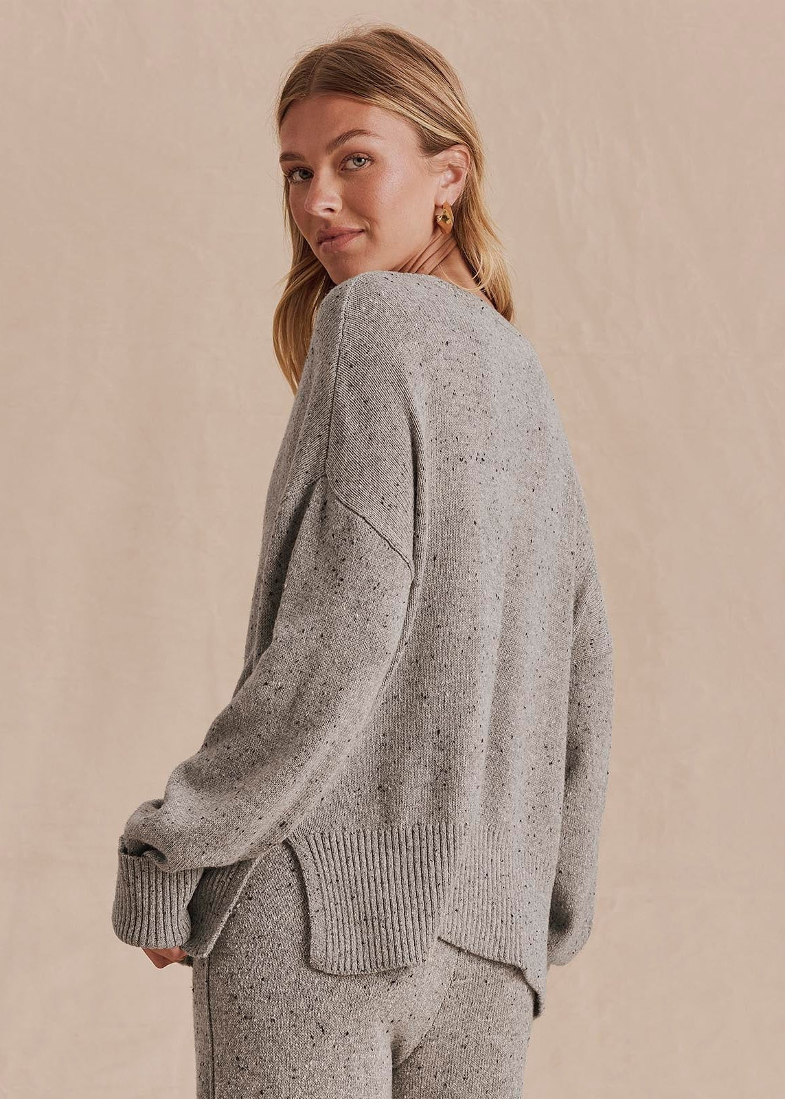 A woman wearing a light gray speckled knit sweater and matching knit pants, photographed against a plain beige studio background.