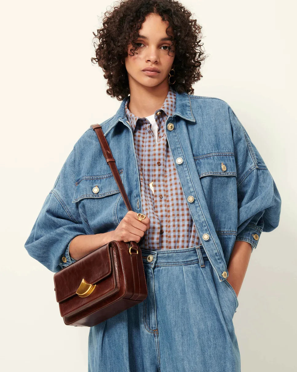 Person wearing a denim jacket and holding a brown leather bag against a plain background