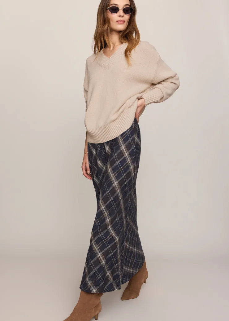 A woman modeling a beige knit V-neck sweater and a navy blue plaid maxi skirt with brown suede ankle boots, photographed against a neutral light gray studio background.