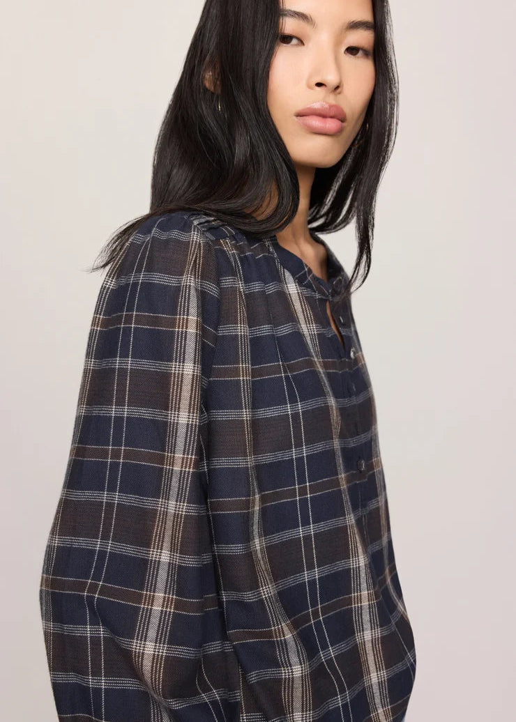 A woman wearing a navy blue plaid button-up shirt with brown and beige stripes, photographed against a neutral light gray background.