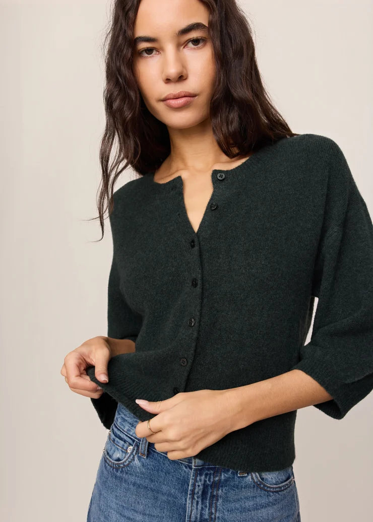 A woman wearing a dark forest green knit cardigan with a split neckline and button front, paired with blue jeans, against a plain beige studio background.