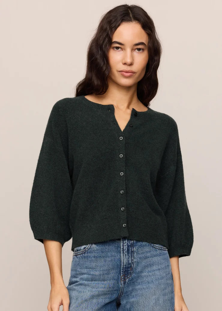 A woman wearing a dark forest green knit cardigan with a buttoned split neckline and three-quarter sleeves, paired with blue jeans, against a solid light beige background.