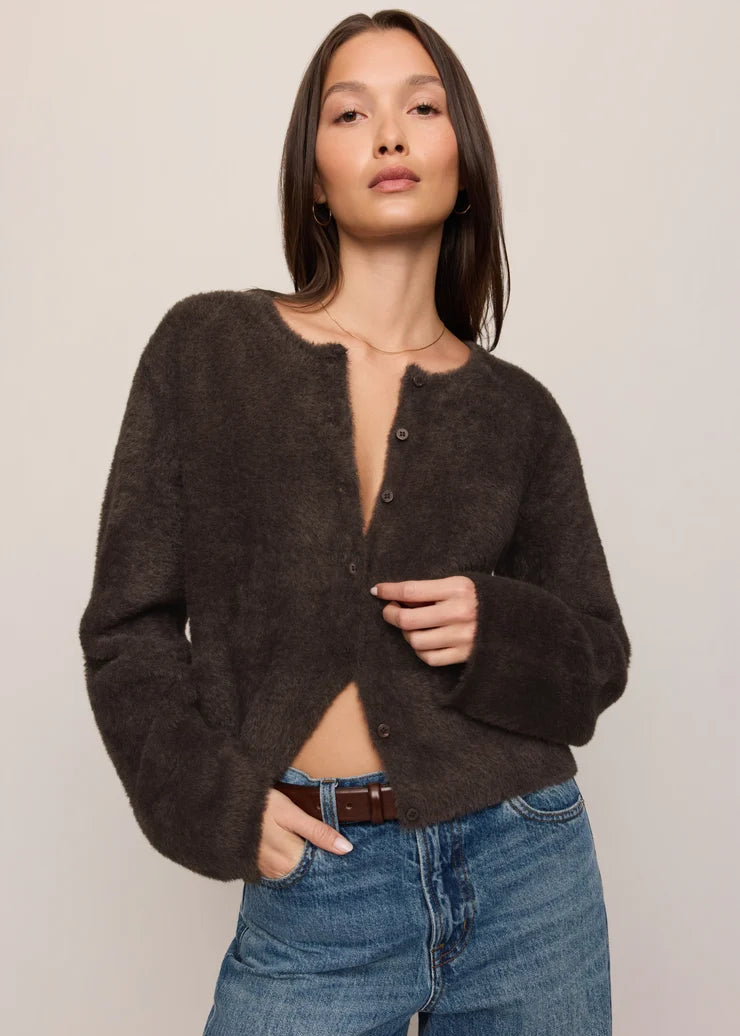 A woman modeling a dark brown fuzzy cropped cardigan with a button front, paired with blue jeans, against a plain light beige studio background.