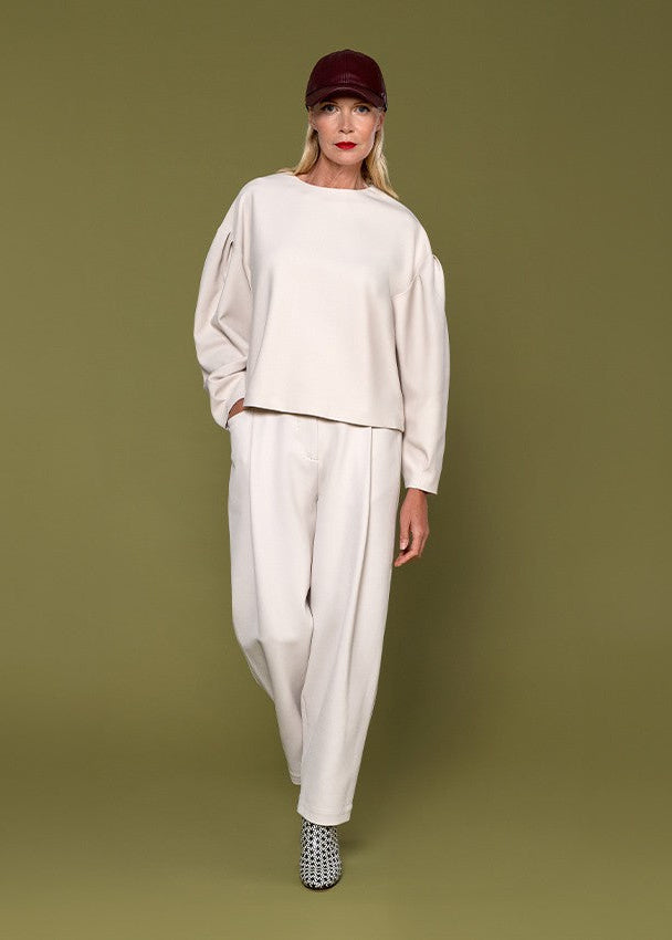 A model stands against a solid olive green studio background wearing a cream-colored two-piece lounge set consisting of a cropped long-sleeve top and matching wide-leg pants, paired with a dark burgundy cap and patterned shoes.