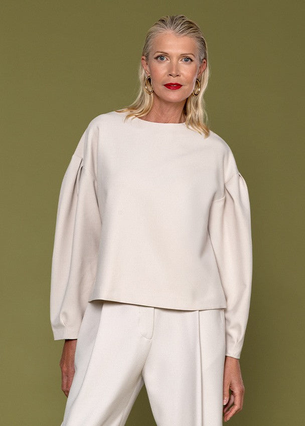 A blonde woman modeling a cream-colored long-sleeve top with dramatic balloon sleeves and matching cream trousers, set against a solid olive green studio background.