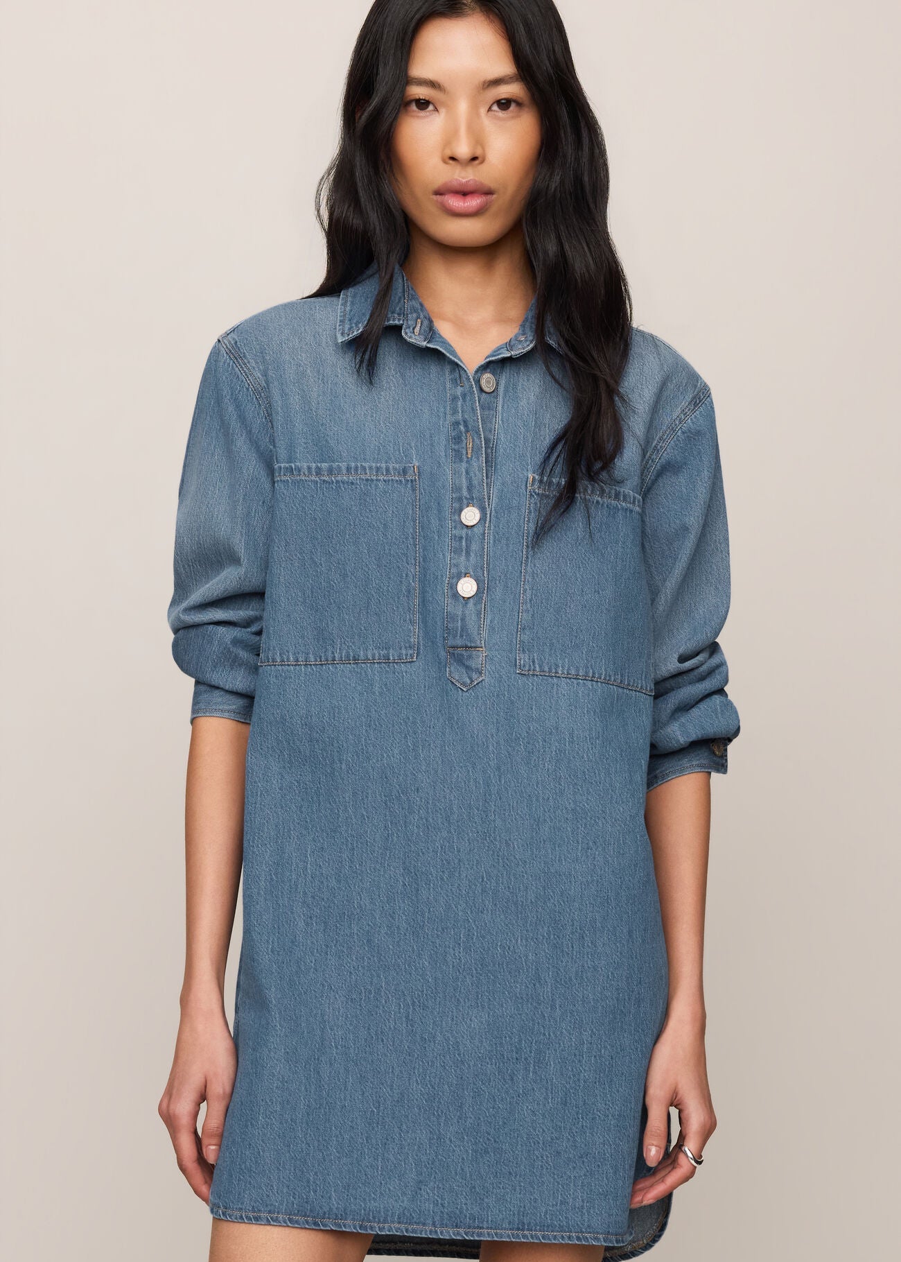 A woman modeling a medium-wash blue denim shirt dress with three-quarter sleeves, a buttoned collar, and two chest pockets, set against a plain beige background.