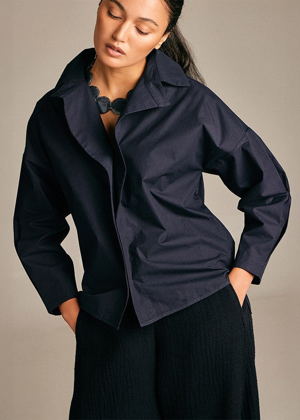 A woman wearing a dark navy blue utility-style jacket with a high collar and front pockets, paired with dark ribbed trousers, photographed against a plain warm gray studio background.