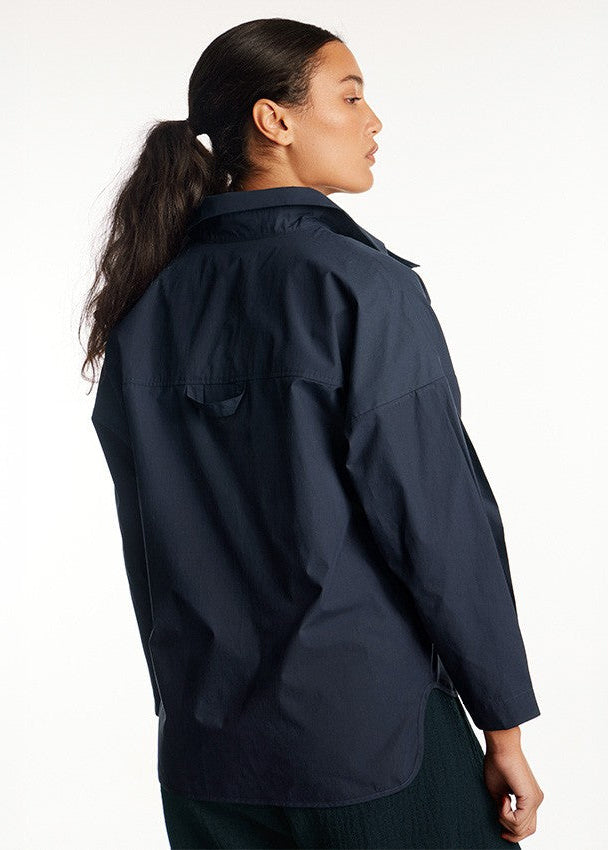 A woman with her hair in a ponytail is shown from the back wearing a dark navy blue utility-style jacket against a plain white studio background.