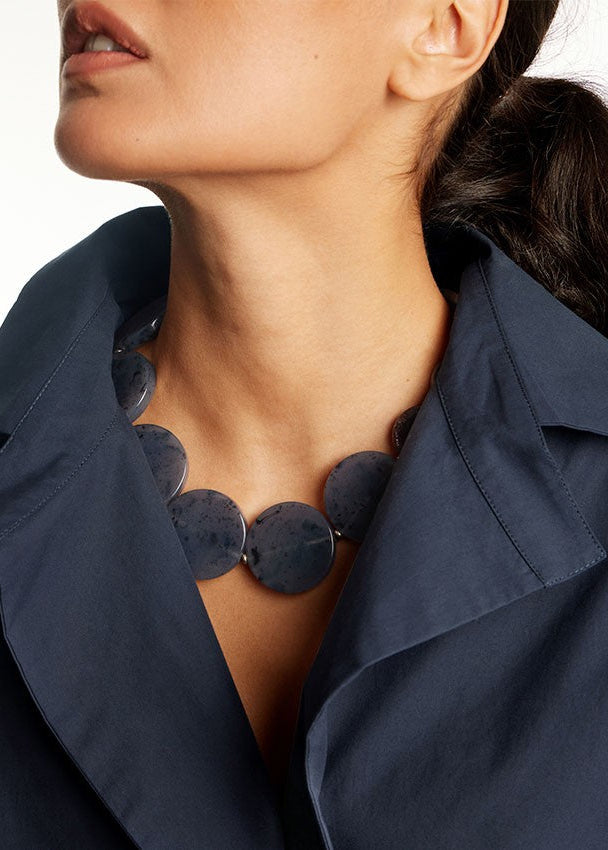 A close-up of a model wearing a chunky statement necklace made of large, dark gray circular discs, styled with a navy blue collared shirt against a white background.