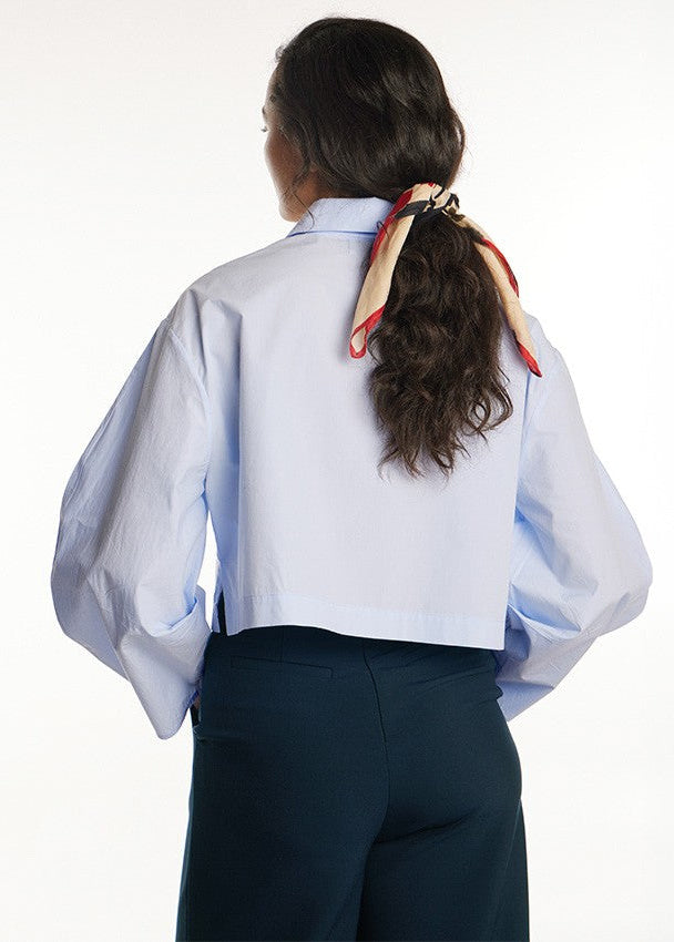 A woman shown from the back wearing a light blue cropped button-up shirt with voluminous sleeves, dark navy trousers, and a red and beige patterned scarf tied in her ponytail.
