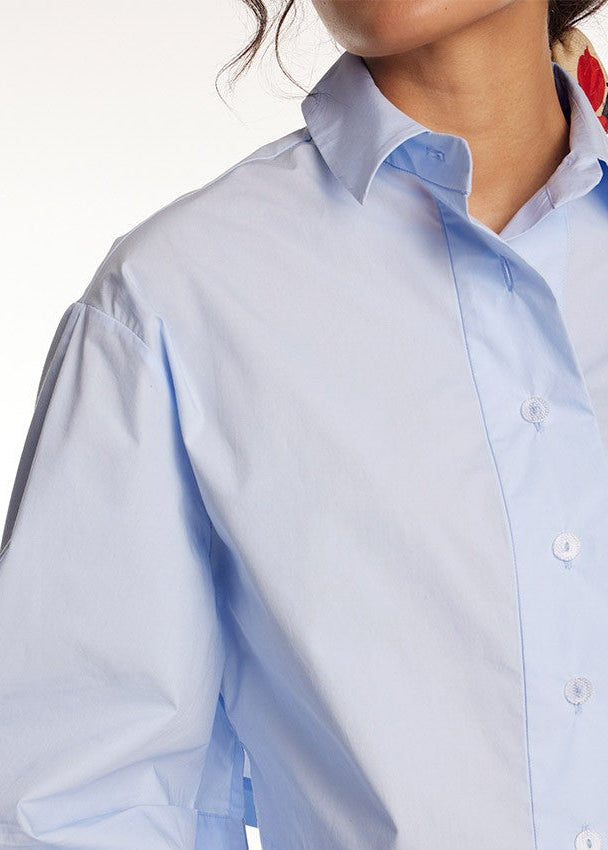 A close-up of a person wearing a light blue long-sleeve button-up shirt against a plain white background.