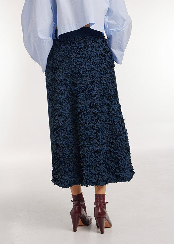 A person shown from the back wearing a dark navy textured midi skirt, a light blue blouse, and dark brown ankle-strap heels against a plain light gray studio background.