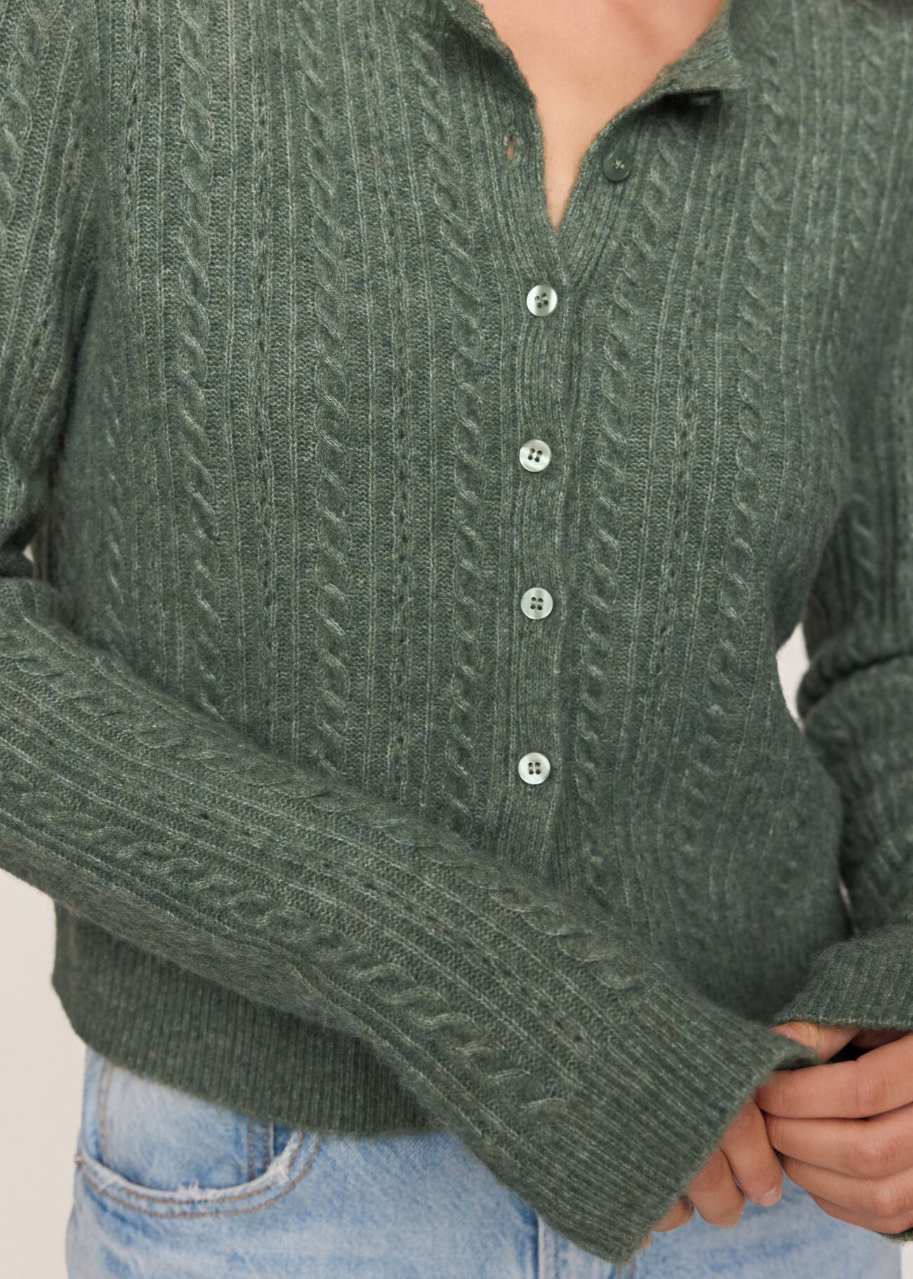 A close-up of a person wearing a textured green cable-knit button-up cardigan sweater with light blue jeans.