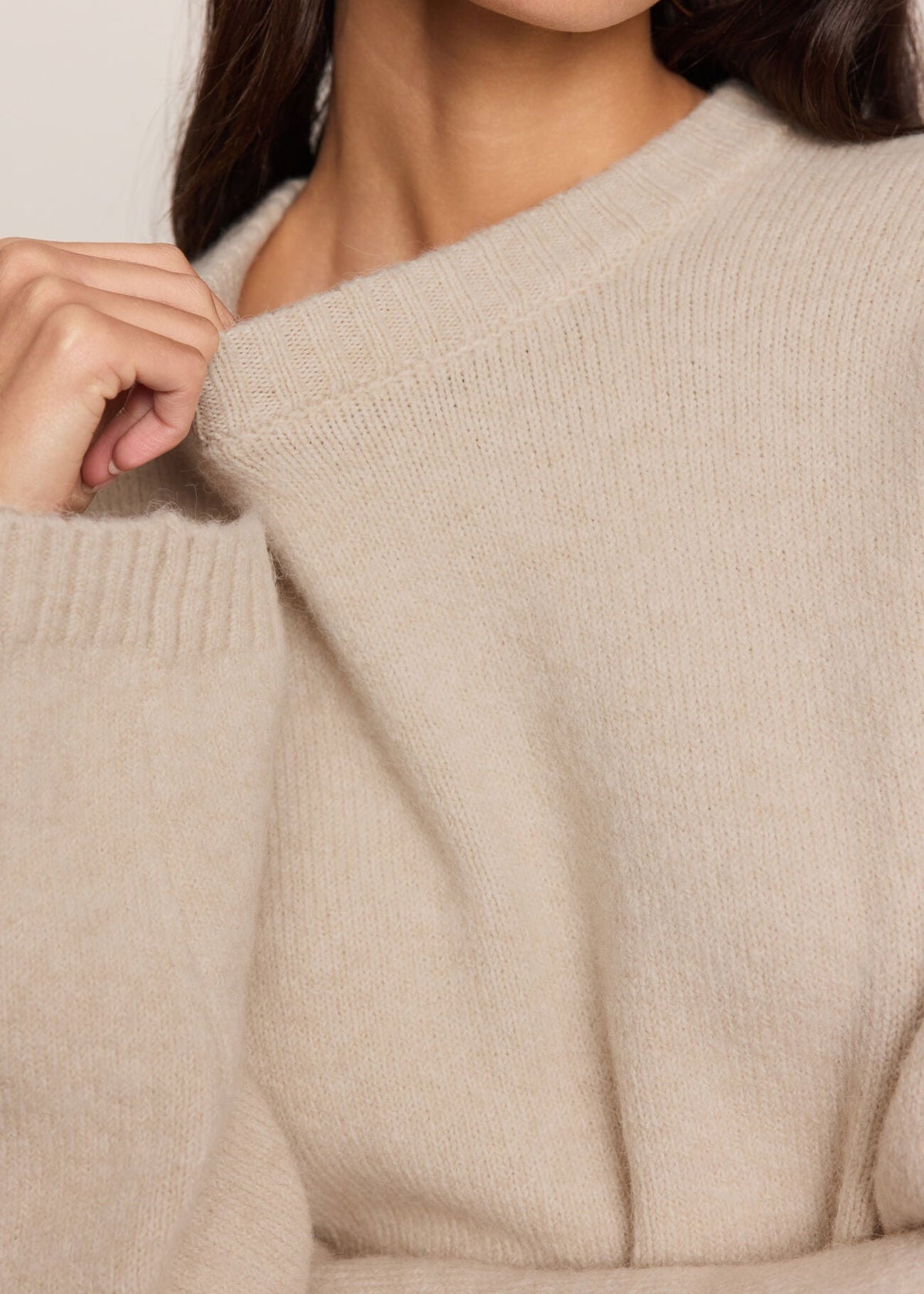 Close-up of a person wearing a soft knit sweater in a light warm beige or oatmeal color, with the hand adjusting the collar.