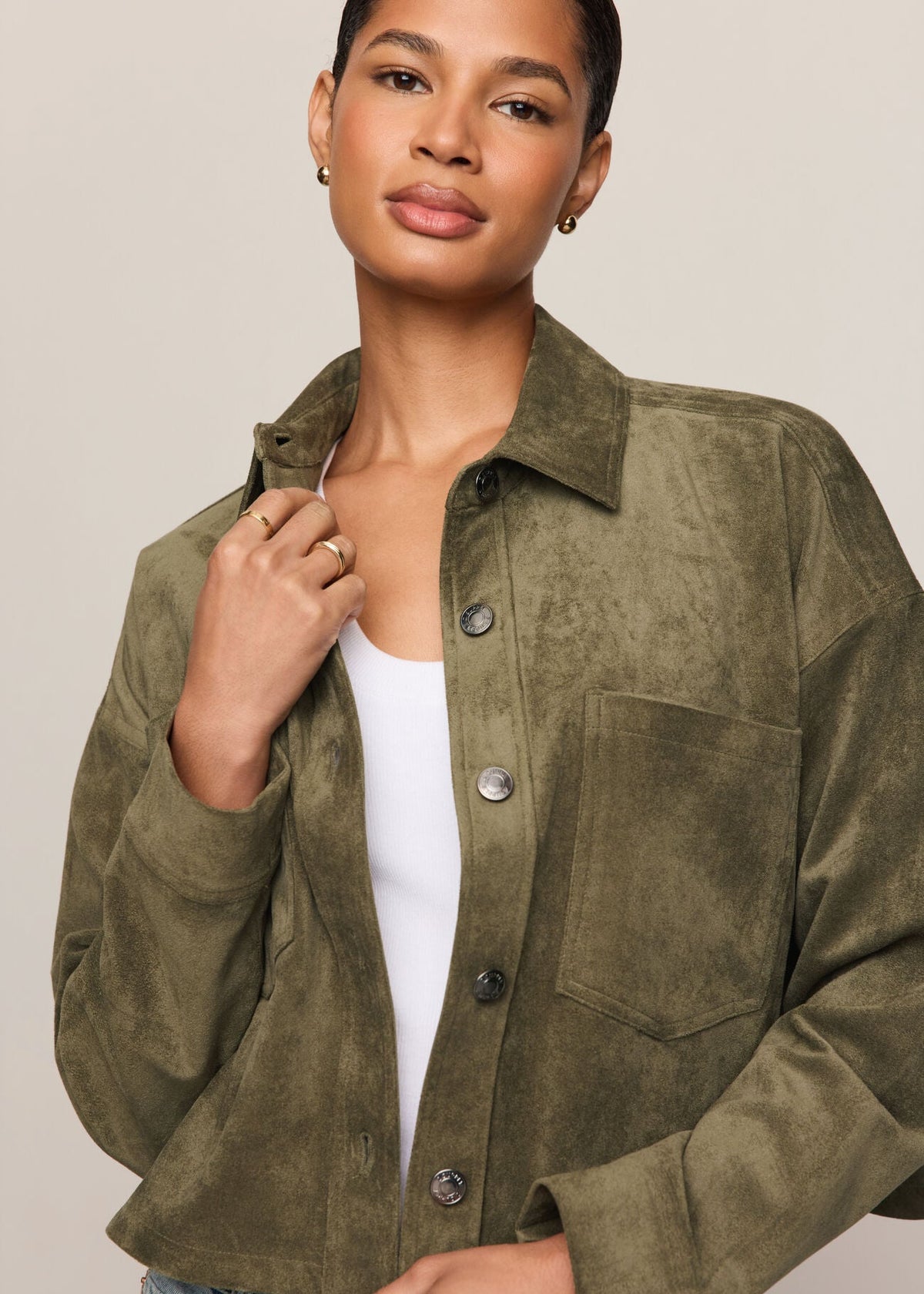 A woman wearing an olive green suede button-up shirt jacket over a white tank top and blue jeans, photographed against a plain light beige studio background.