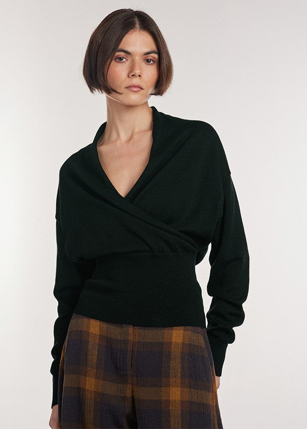 A model with a short dark bob haircut wearing a dark forest green wrap-style knit sweater and brown and navy plaid trousers, photographed against a light gray studio background.