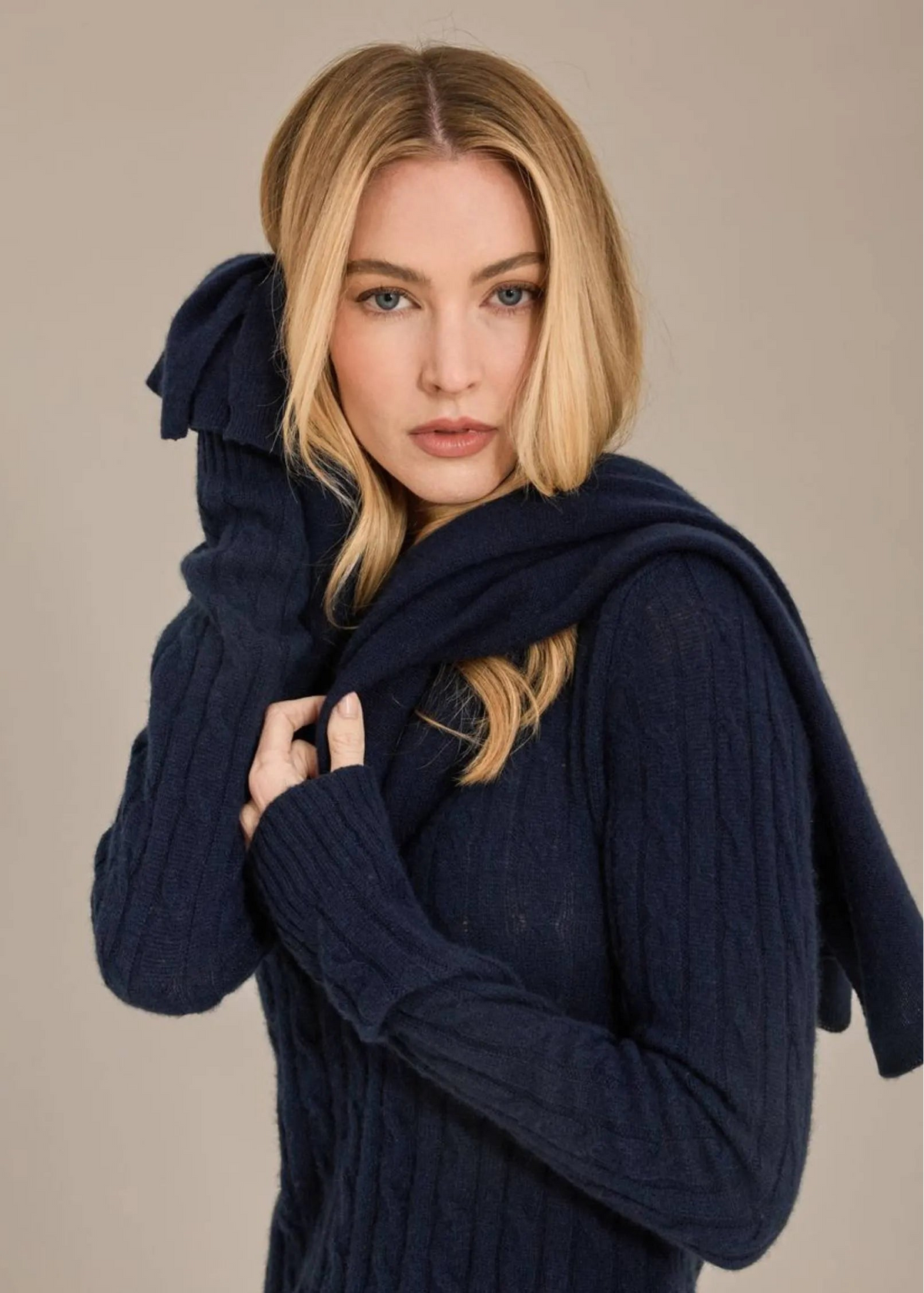A blonde woman wearing a dark navy blue ribbed knit sweater and matching scarf, posing against a solid beige studio background.
