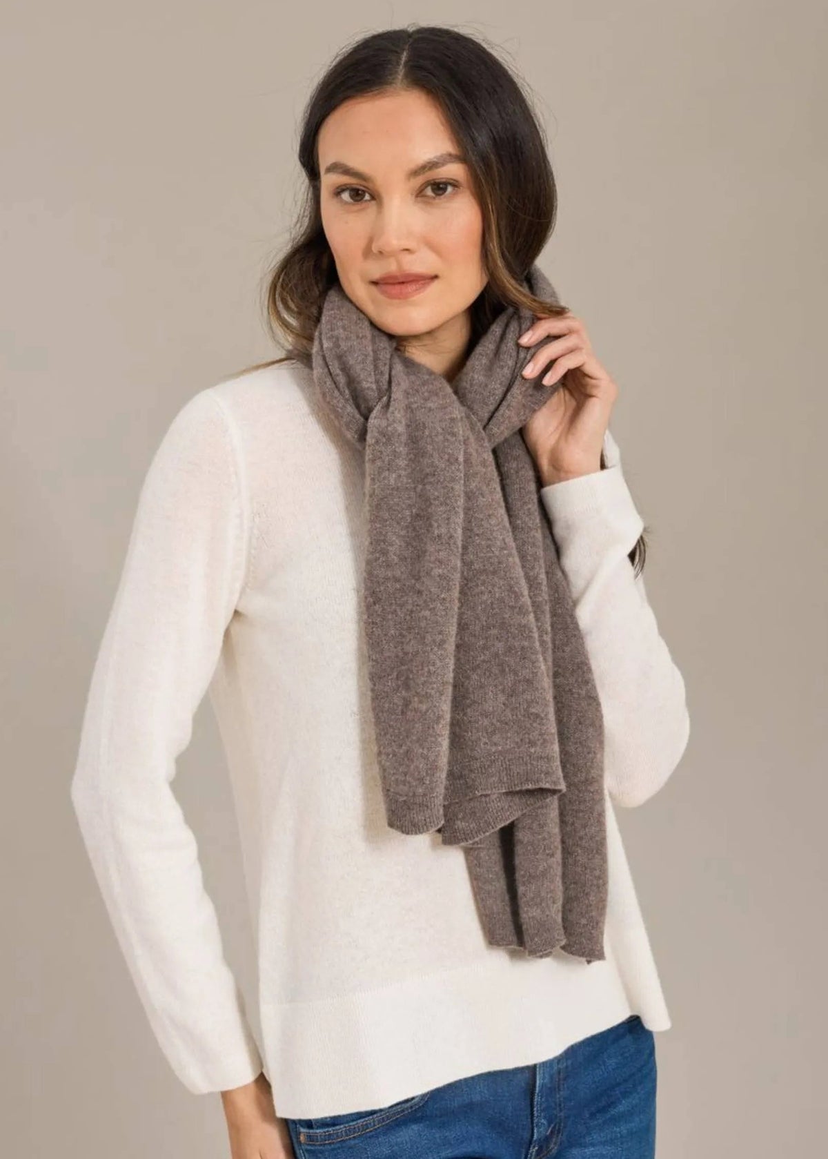 A woman wearing a white knit sweater and blue jeans, wrapped in a heathered taupe-brown scarf, posed against a solid light gray studio background.