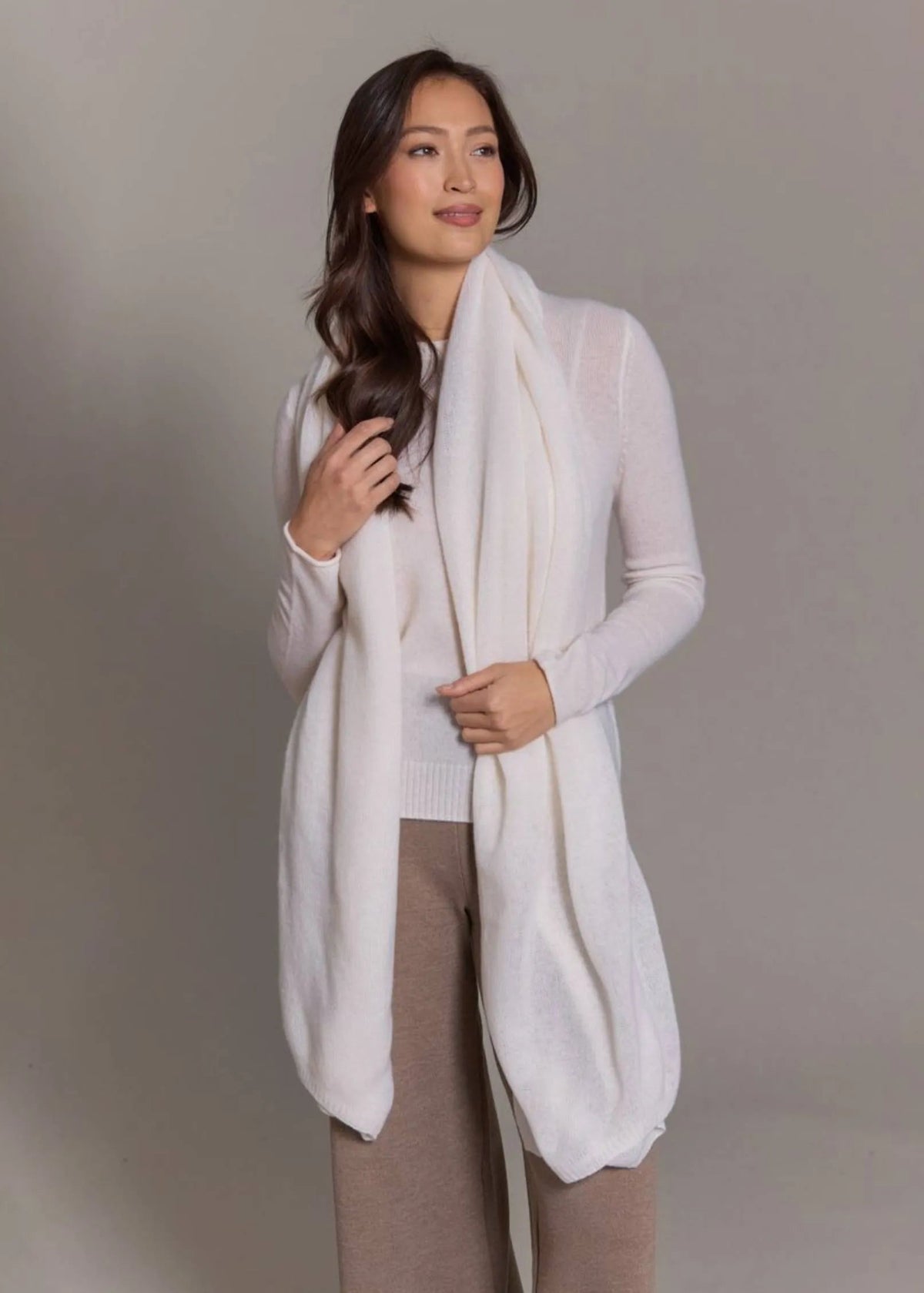 A woman modeling a long, lightweight cream knit cardigan with a matching draped scarf, paired with taupe pants, against a neutral gray studio background.