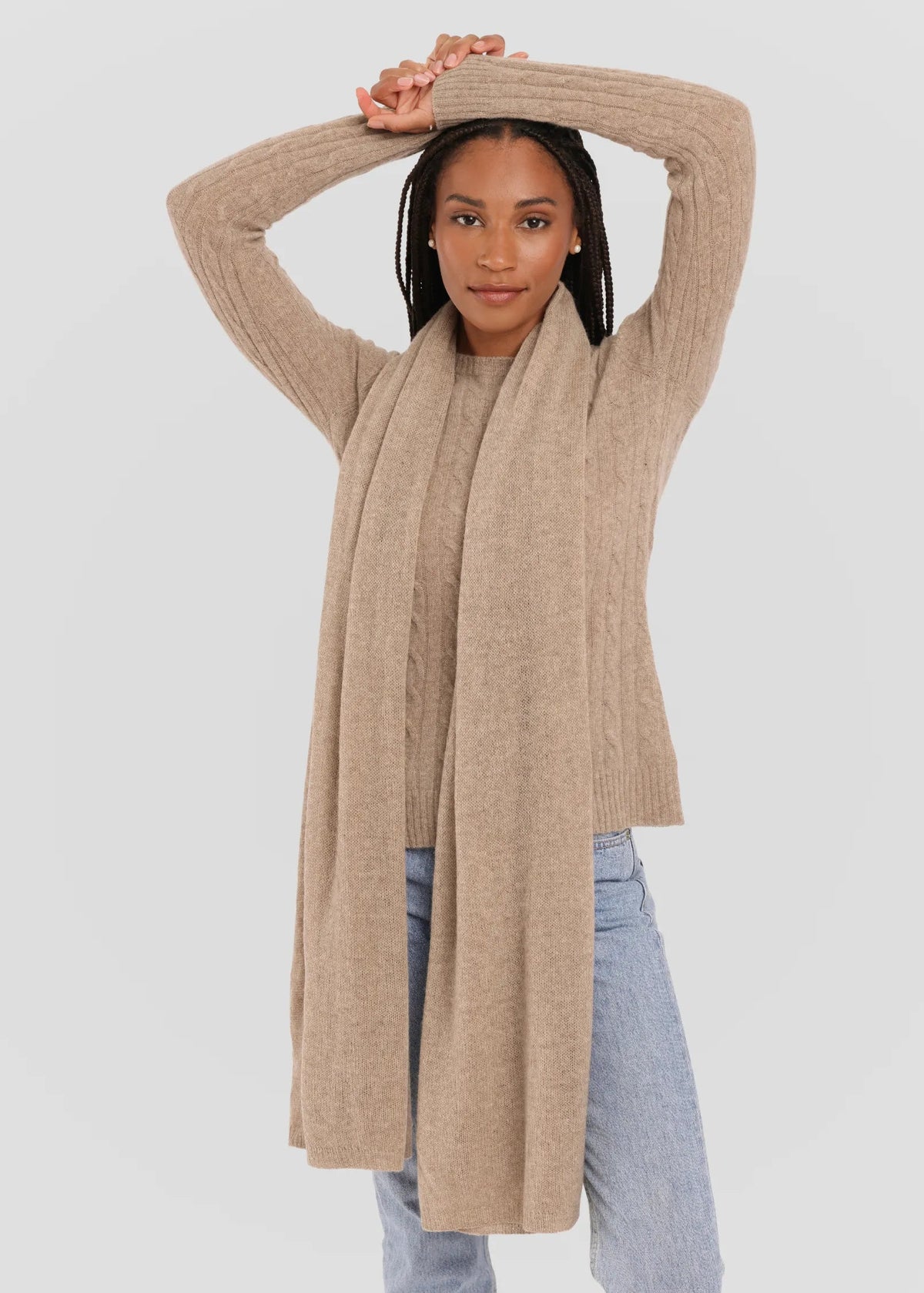 A woman modeling a long, light beige ribbed knit cardigan sweater with a matching knit scarf, paired with light blue jeans, against a solid light gray background.